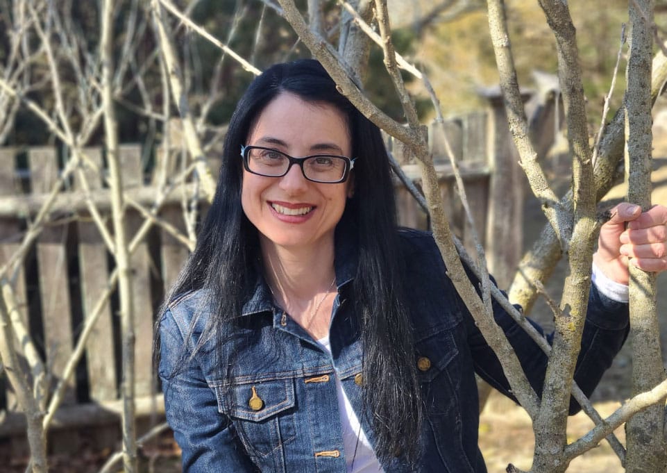 Voula Manousos, weight management and pregnancy nutritionist, smiles at the camera in a denim jacket, as she rests next to a small tree