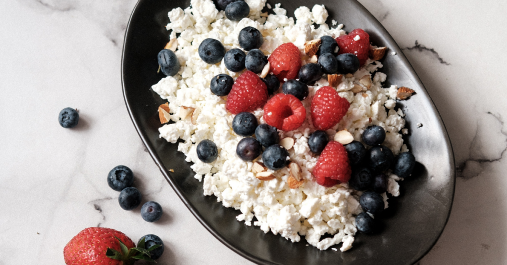 A complete and healthy snack option, consisting of a bowl of cottage cheese topped with fresh blueberries and raspberries, as well as chopped almonds