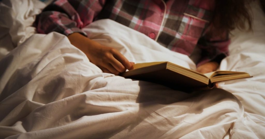 A woman's hands hold a book propped up on wrinkles of fabric in bed