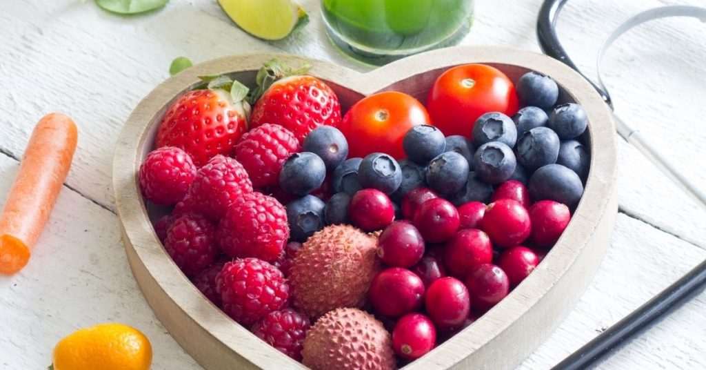 A heart-shaped bowl sits on a table filled with berries, fruits, and vegetables
