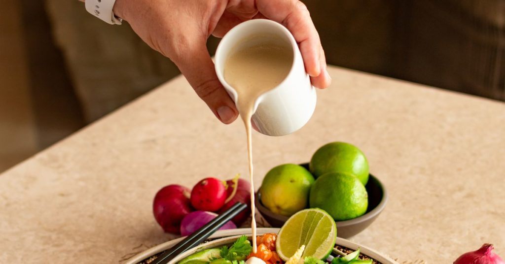 A hand holds up a small carafe pouring out salad dressing onto a fresh green salad