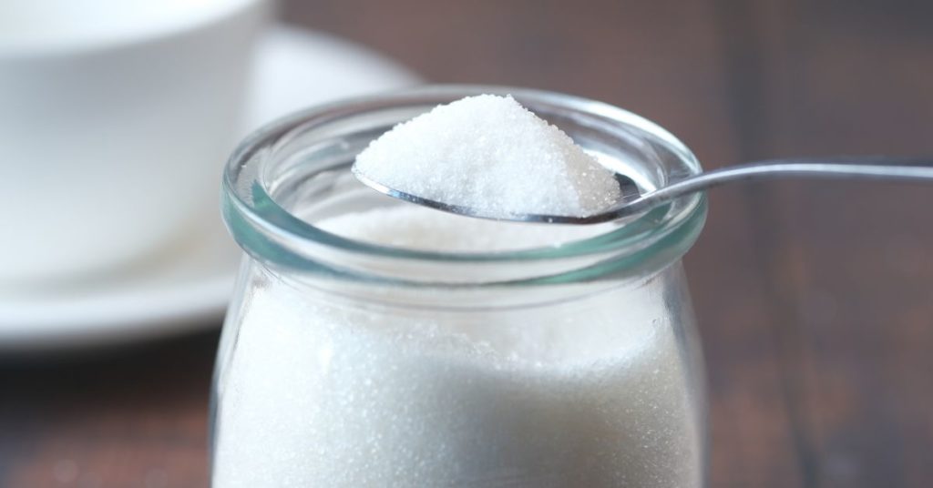 A jar full of sugar with a full teaspoon being taken out of it