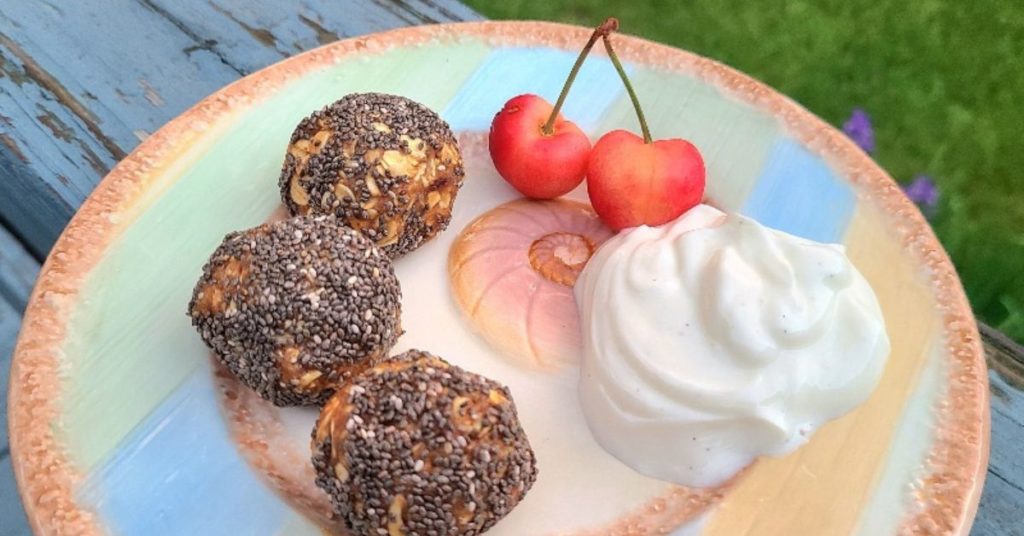 3 peanut butter balls dusted in chia seeds are set on a plate next to yogurt and cherries
