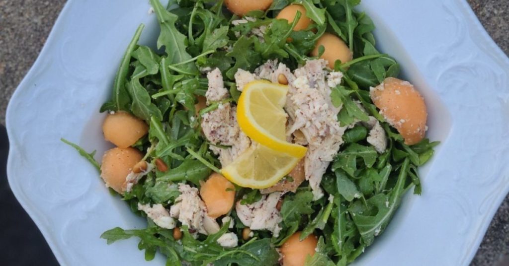 Arugula salad topped with balls of cantoloupe melon, shredded chicken and decorated with a lemon twist