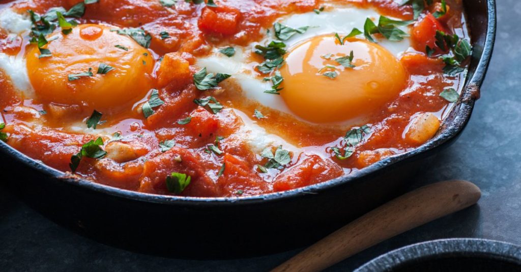 Eggs cooking in an inch deep of tomato sauce inside a cast iron pan and garnished with fresh herbs