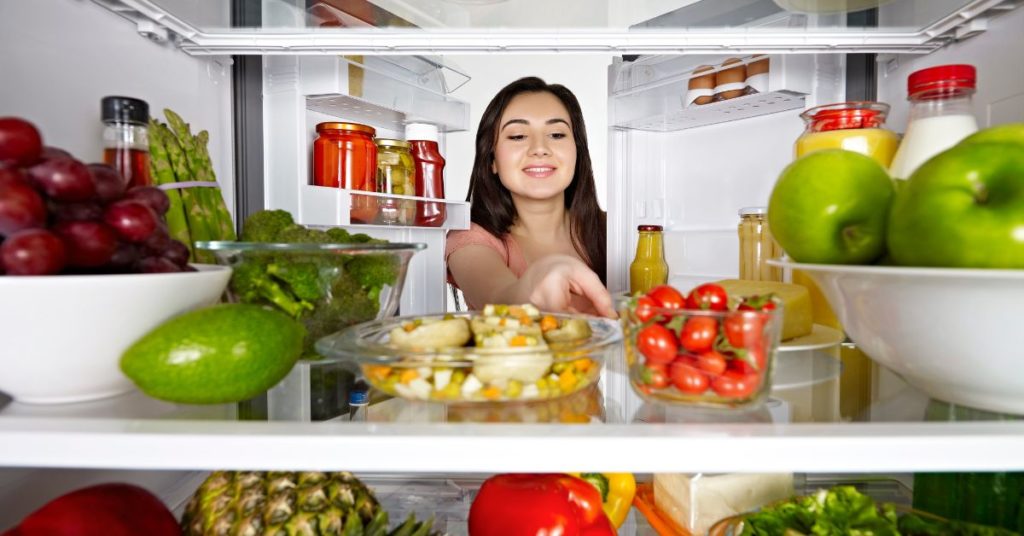 Clean Eating Recipes for Beginners, view from inside the fridge of a woman opening fridge doors and reaching in to grab fresh vegetables. Fridge is stocked full of clean eating healthy foods.