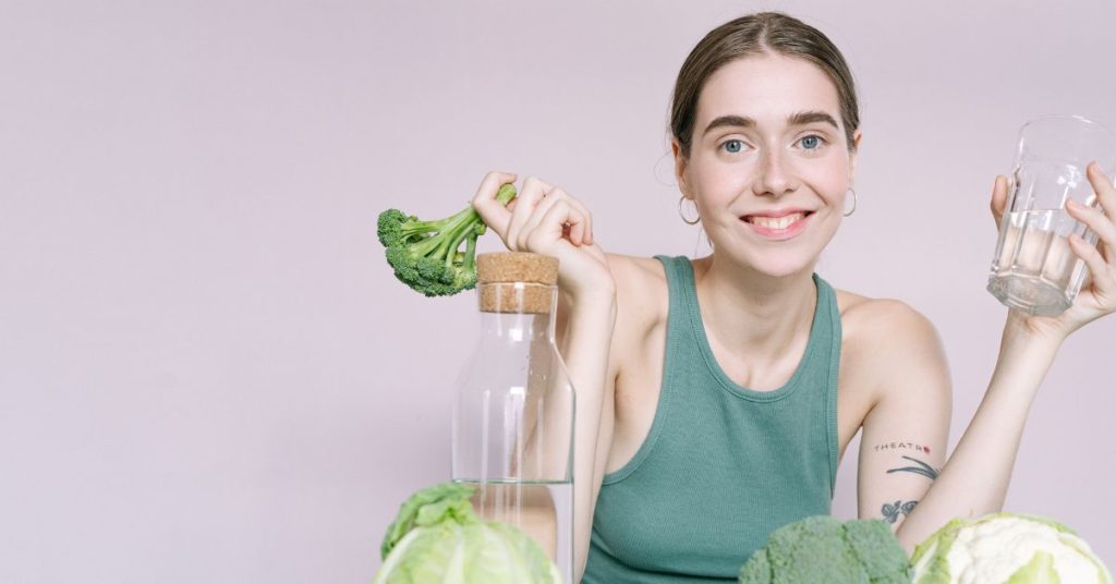 A woman looks holds her hands up in a shrug, with water and vegetables in front of her