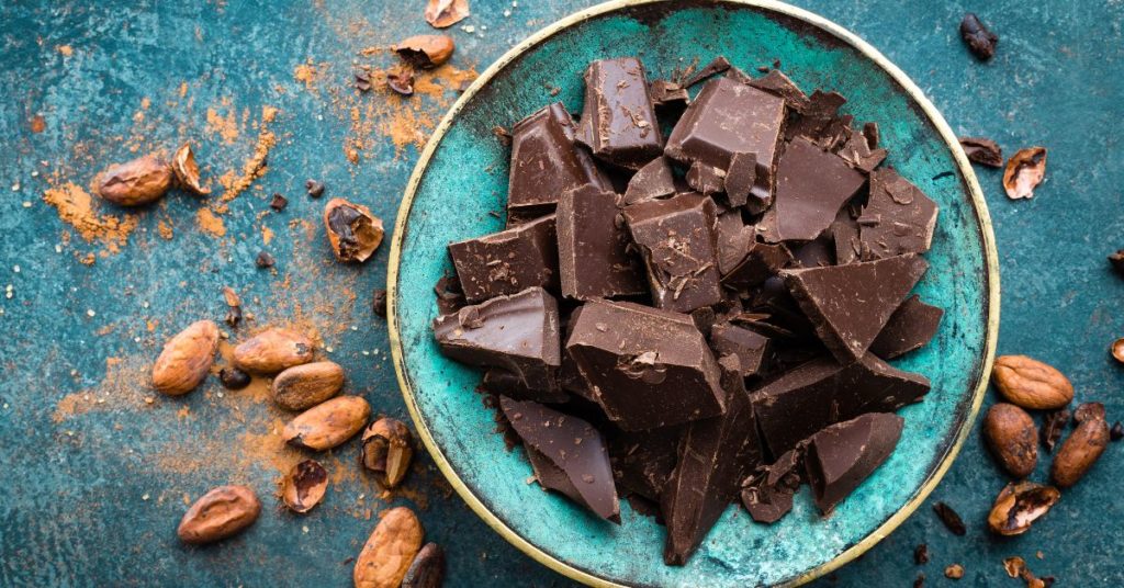 A bowl of dark chocolate chunks is surrounded by fresh cacao beans