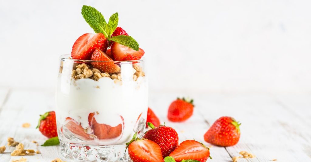 A transparent glass holds Greek yogurt topped with fresh strawberries, mint, and granola