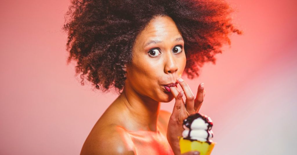A woman looks at the camera wide-eyed as if she was caught, while she holds a chocolate-drizzled ice cream cone and licks her finger, Ways to Stop Sugar Craving