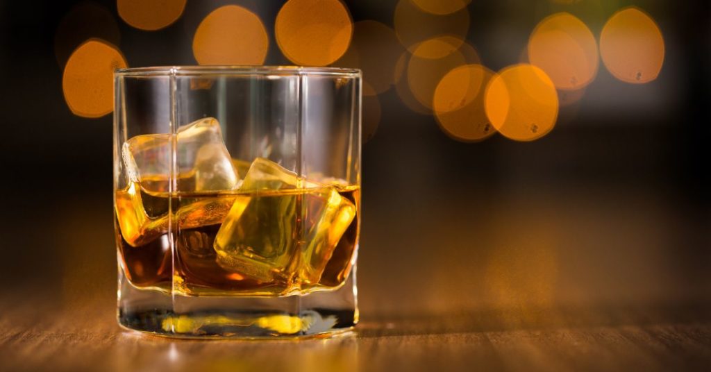 A glass of scotch on the rocks set on a wooden tabletop with blurred holiday lights in the background | Alcohol and Weight Gain During Holidays