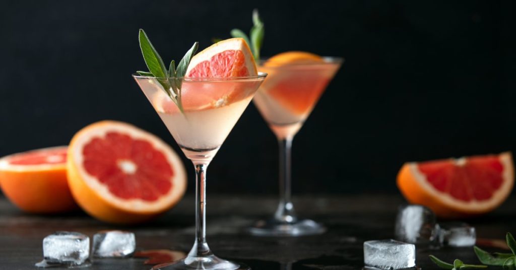 A martini glass filled with a mocktail consisting of a light liquid and fresh grapefruit and sage