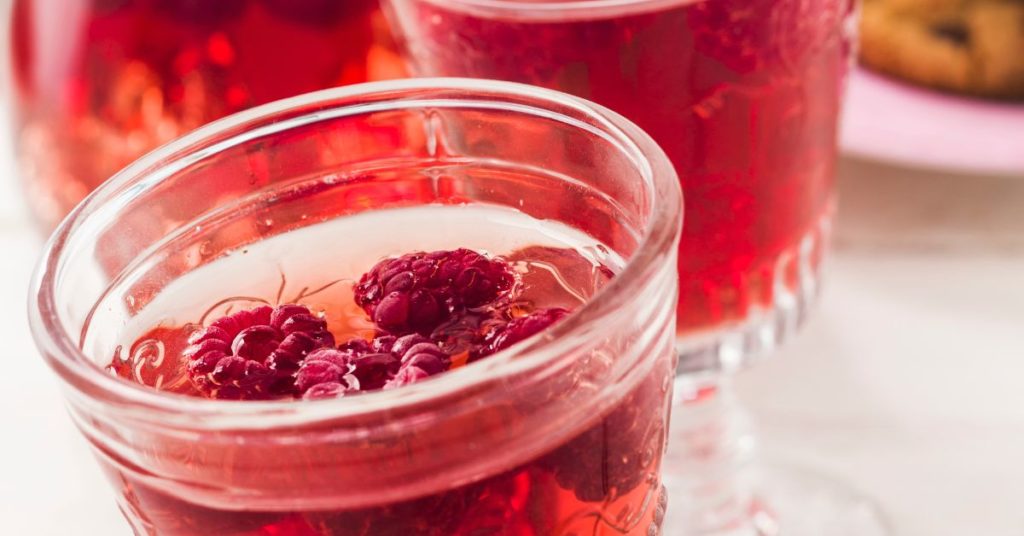 A mocktail served in a mason jar with a clear, colorful liquid and raspberries floating on top