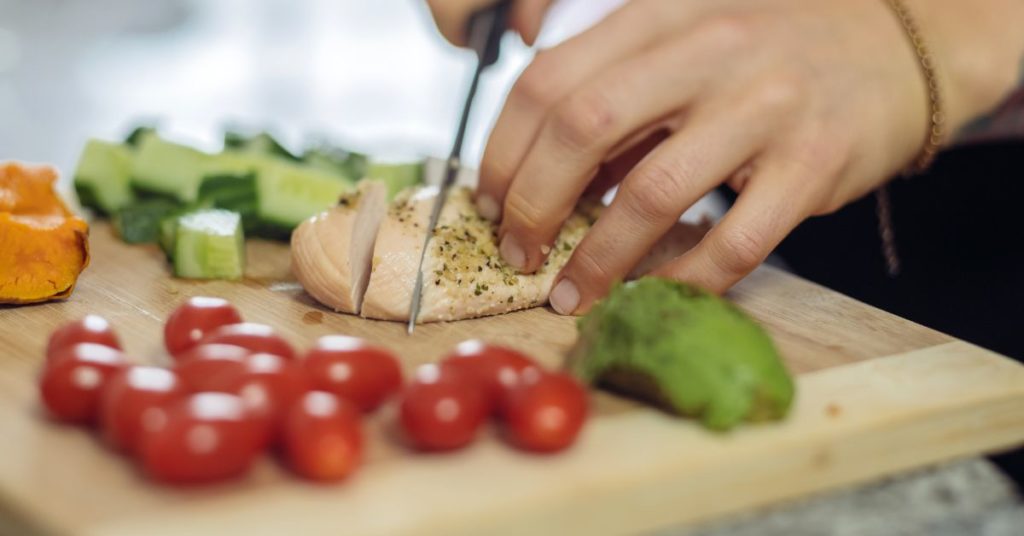 A woman slices a cooked chicken breast as she meal preps with lots of veggies next to her on her cutting board