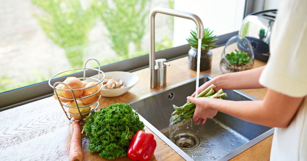 A woman washes asparagus in her kitchen sink with more vegetables ready to be washed, cut and prepped sit on the counter next to her