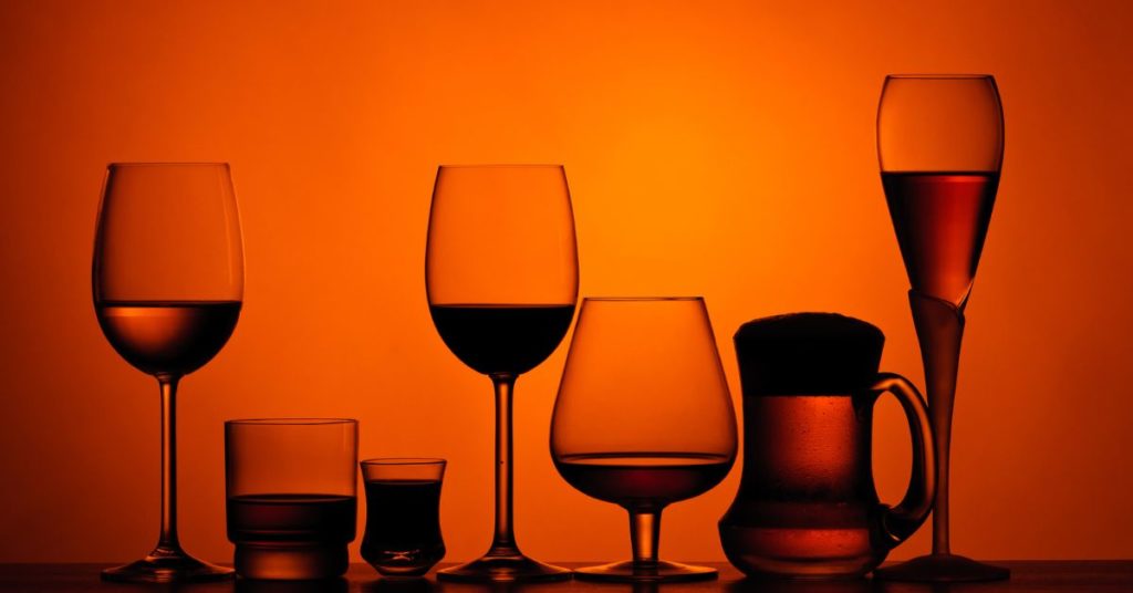 Different types of alcohol glasses filled with alcohol set in a row and backlit by a moody, dark light | Alcohol and Weight Gain During Holidays