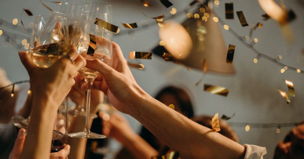 Multiple people hold up glasses of alcohol at a party with golden confetti shimmering down around them | Alcohol and Weight Gain During Holidays