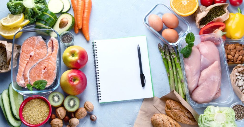 Several fresh foods like eggs, salmon, chicken, fruit, and vegetables surround a notebook and pen for meal planning and meal prep for healthy weight loss