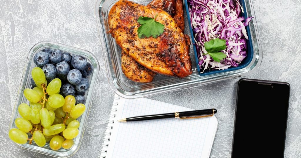 Two glass containers of prepared food filled with roast chicken, cabbage slaw, grapes and blueberries on a counter next to a open notebook and cell phone, Meal Prep Ideas for Weight Loss