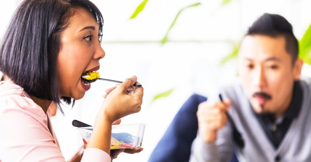 A woman shovels food into her mouth from a plastic container on what looks to be a lunch break, mindful eating for weight loss