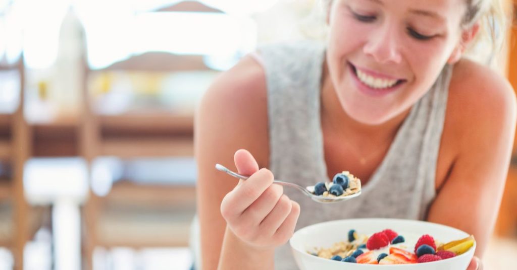 A young happy woman holds a bowel of cereal topped with fresh berries, she's taking a spoonful of it and is mindfully considering what she is eating, mindful eating for weight loss