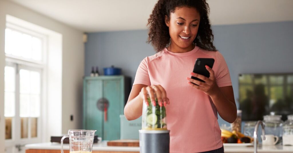 A woman looks at her phone as she makes a smoothie, as an example of how you can use apps and tracking for SMART goals for weight loss