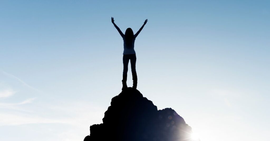 The silhouette of a person stands atop a mountain with arms widespread in celebration of achieving their SMART goal