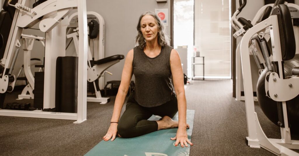 A grey-haired woman sits in pigeon pose on a yoga mat, there is weight machines all around her, menopausal weight gain