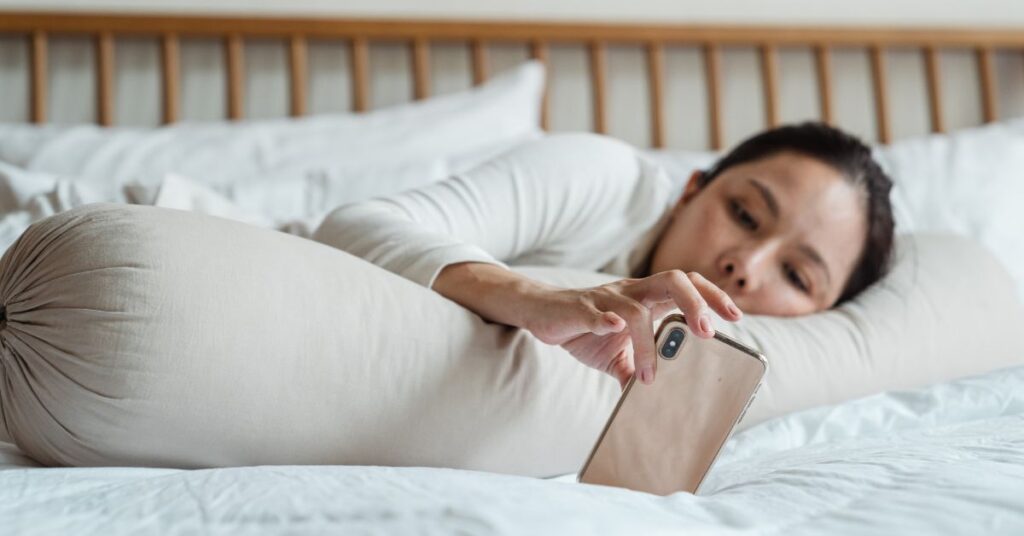 Woman lays in bed and looks at her phone, How to Stop Cortisol Weight Gain