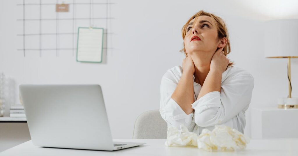 Woman working on a laptop holds onto her shoulders in a stressful pose, How to Stop Cortisol Weight Gain