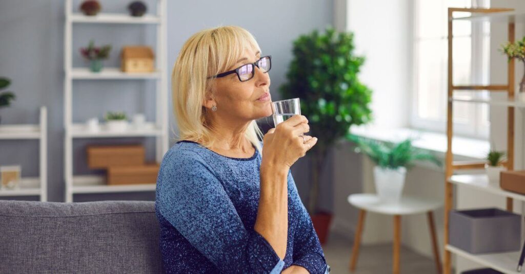 An older blonde woman in glasses sips thoughtfully out of a glass of water, portion control