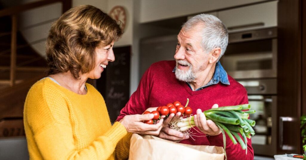 An older couple laughs with each other as they unpack vegetables from a brown paper bag in their kitchen