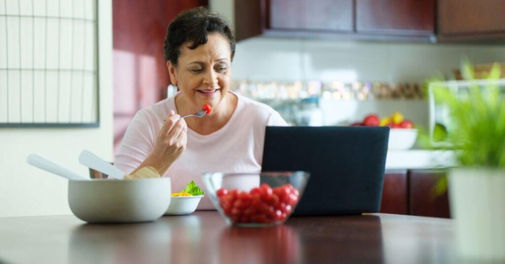 An older woman with short hair eats a cherry tomato out of her salad while watching something on her laptop, portion control