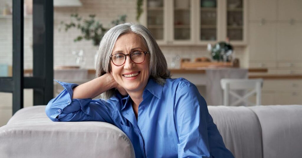 A healthy gray-haired woman in glasses rests on a couch and smiles a toothy grin