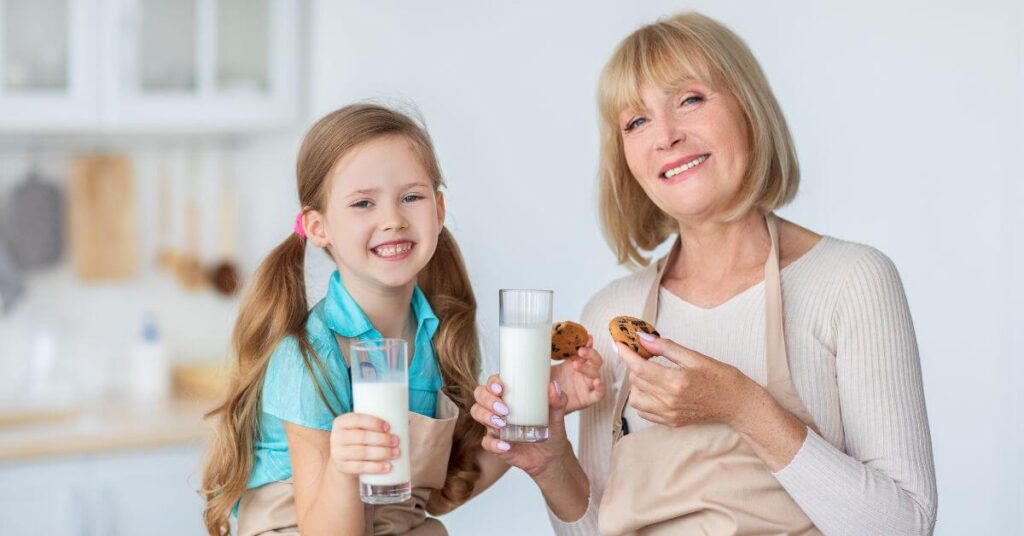 A middle-aged woman and a little girl wearing aprons each hold chocolate chip cookies and glasses of milk