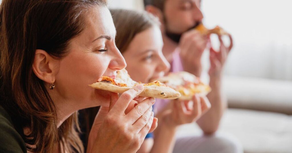 A middle aged woman takes a bite of pizza with her husband and child also eating pizza blurred out in the background