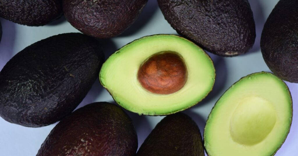 A ripe avocado is sliced in half amongst several whole avocados, healthy fats