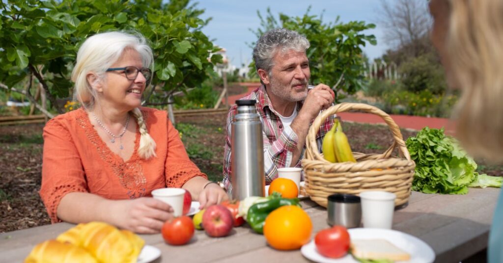 A white-haired woman and man sit at a picnic table with various fruits and veggies spread out around them