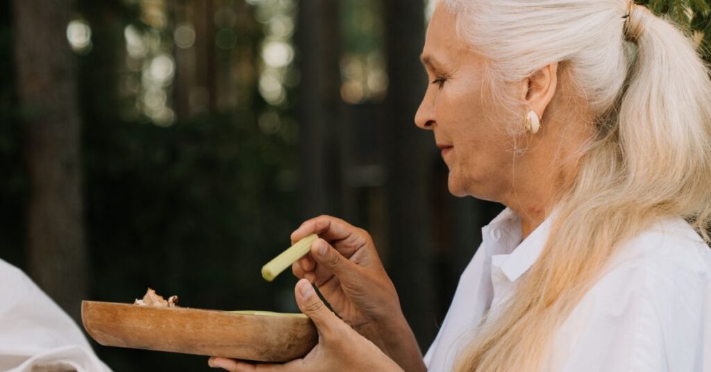 A white-haired woman takes a piece of celery and dips it into a spread