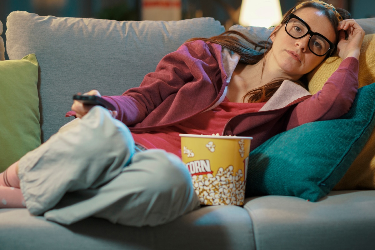 Woman sitting on a couch watching television and eating popcorn, illustrating emotional eating triggered by stress or sadness